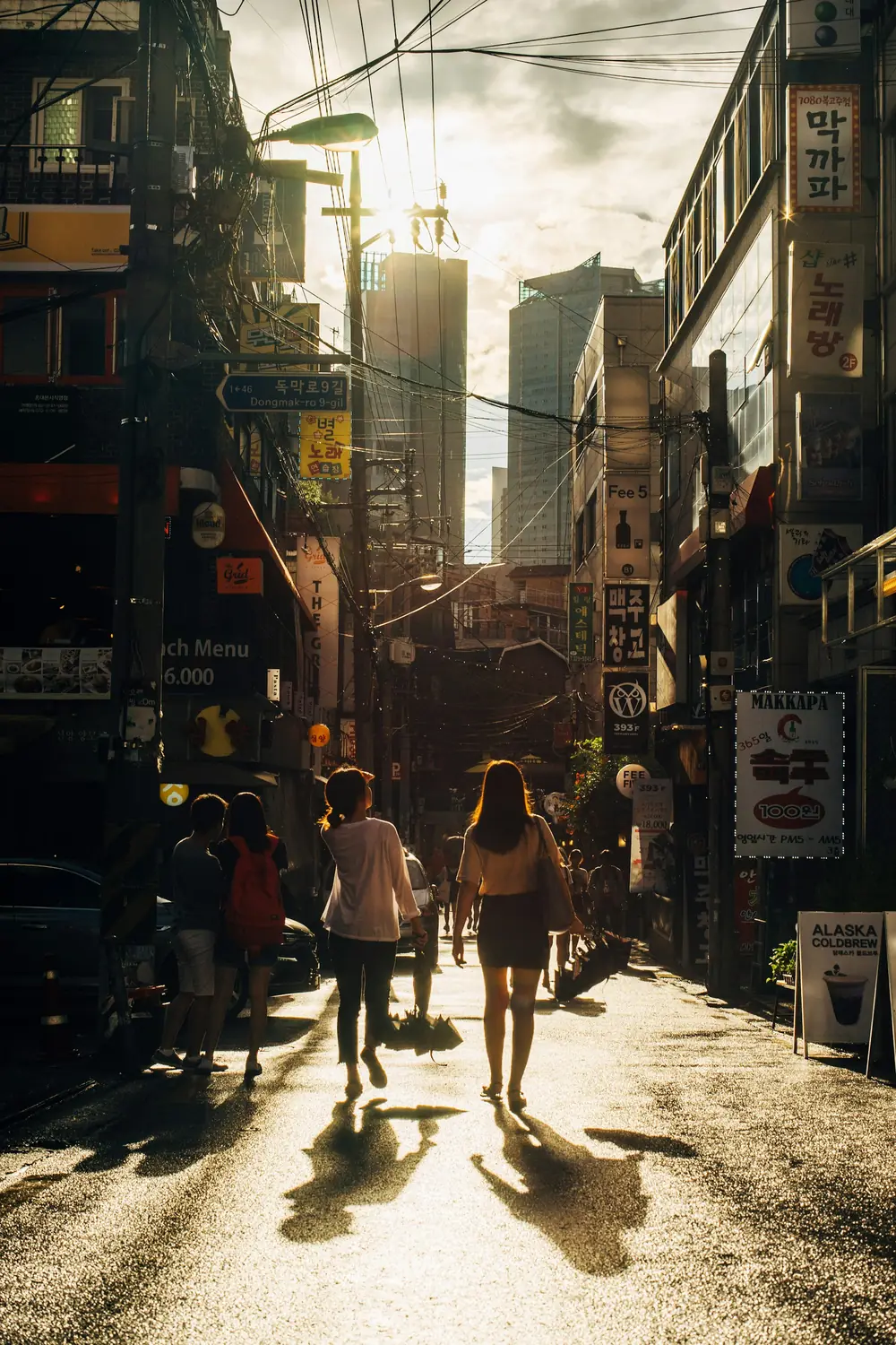 People walking along a narrow Seoul street lined with Korean shop signs at sunset